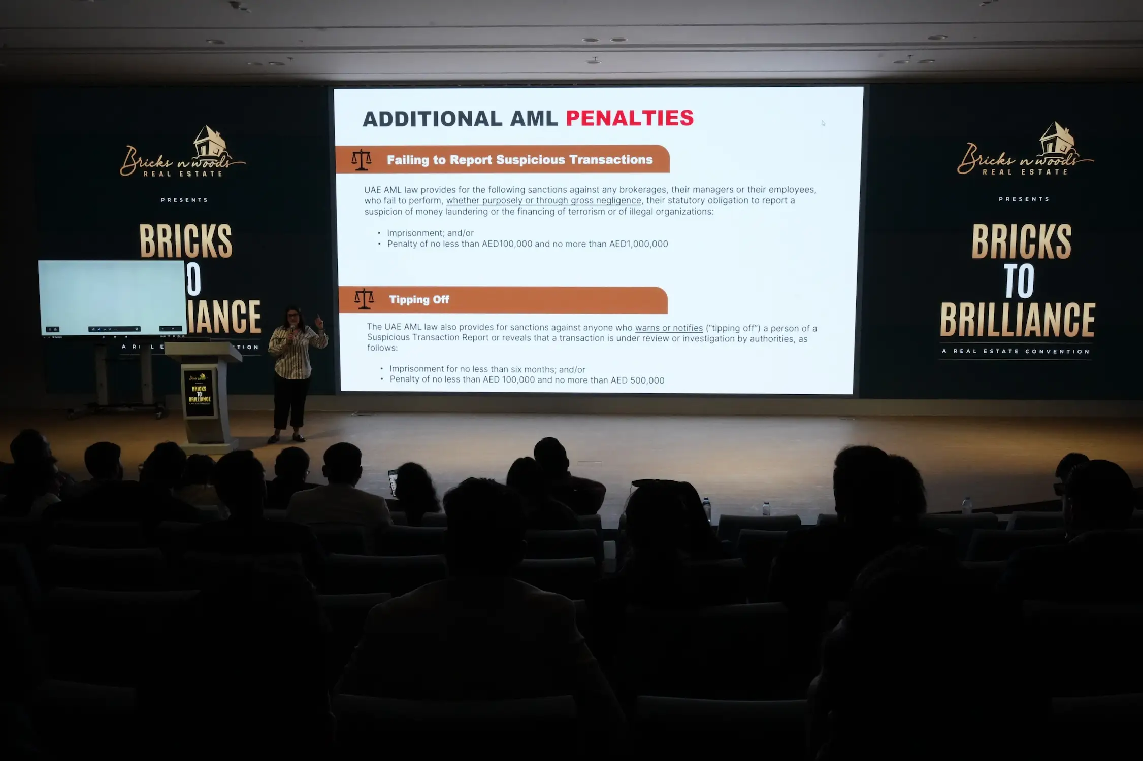 A man presenting at a conference and visuals on an LED screen advertising behind him.