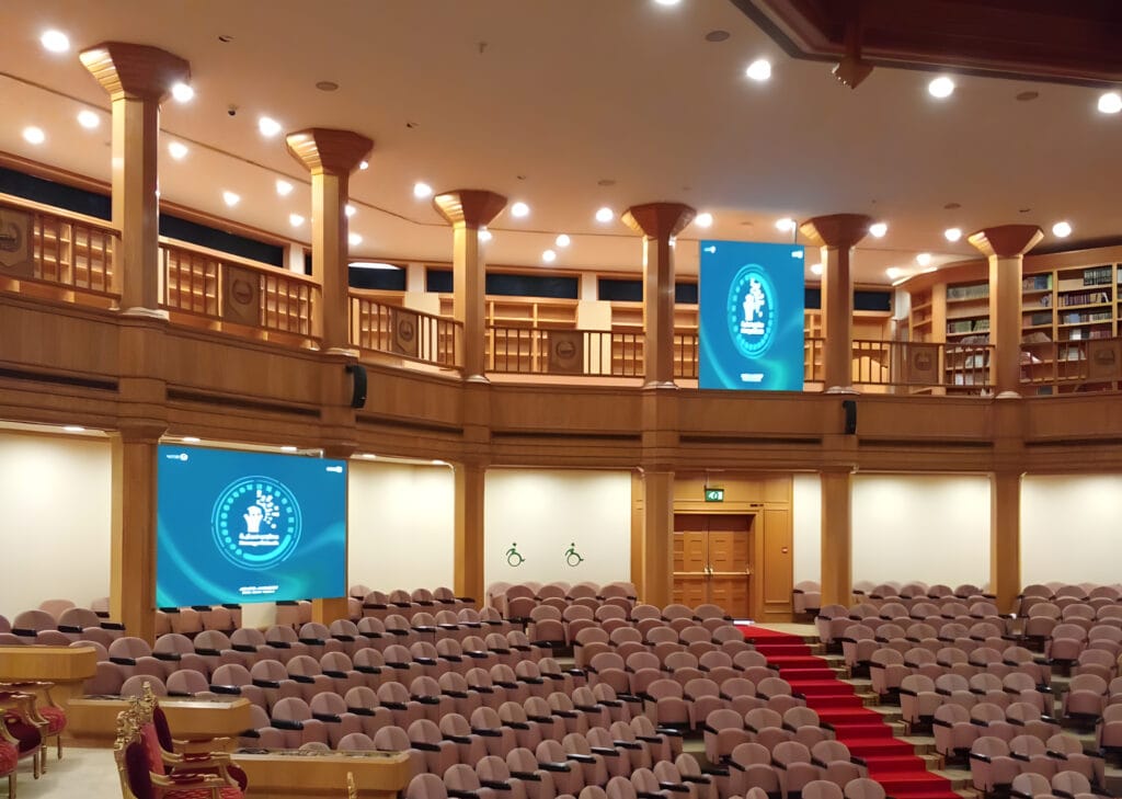 An auditorium featuring chairs and a stage, with indoor screens positioned for presentations.